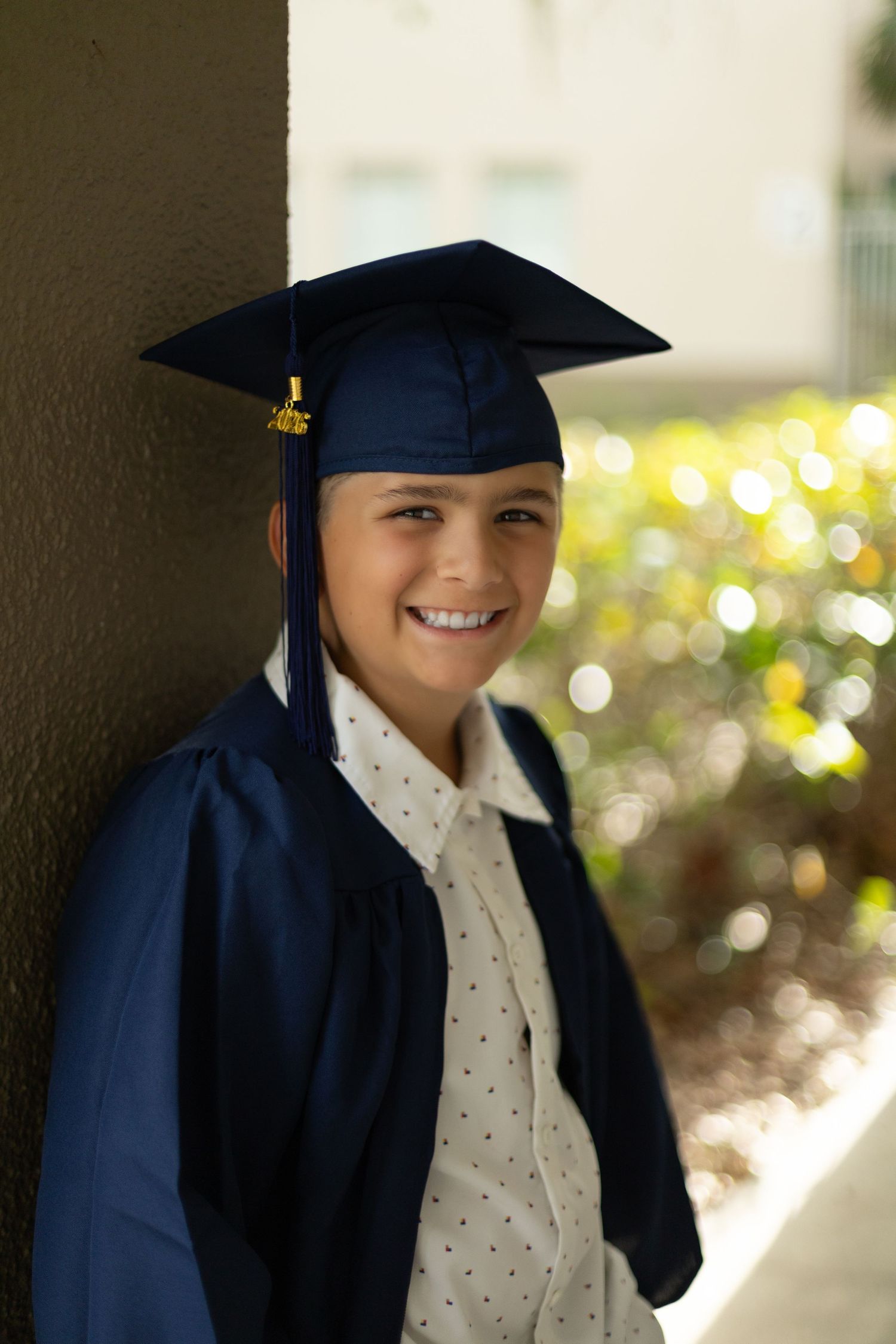 Smiling graduate in navy blue cap and gown leaning against a column outdoors on graduation day.