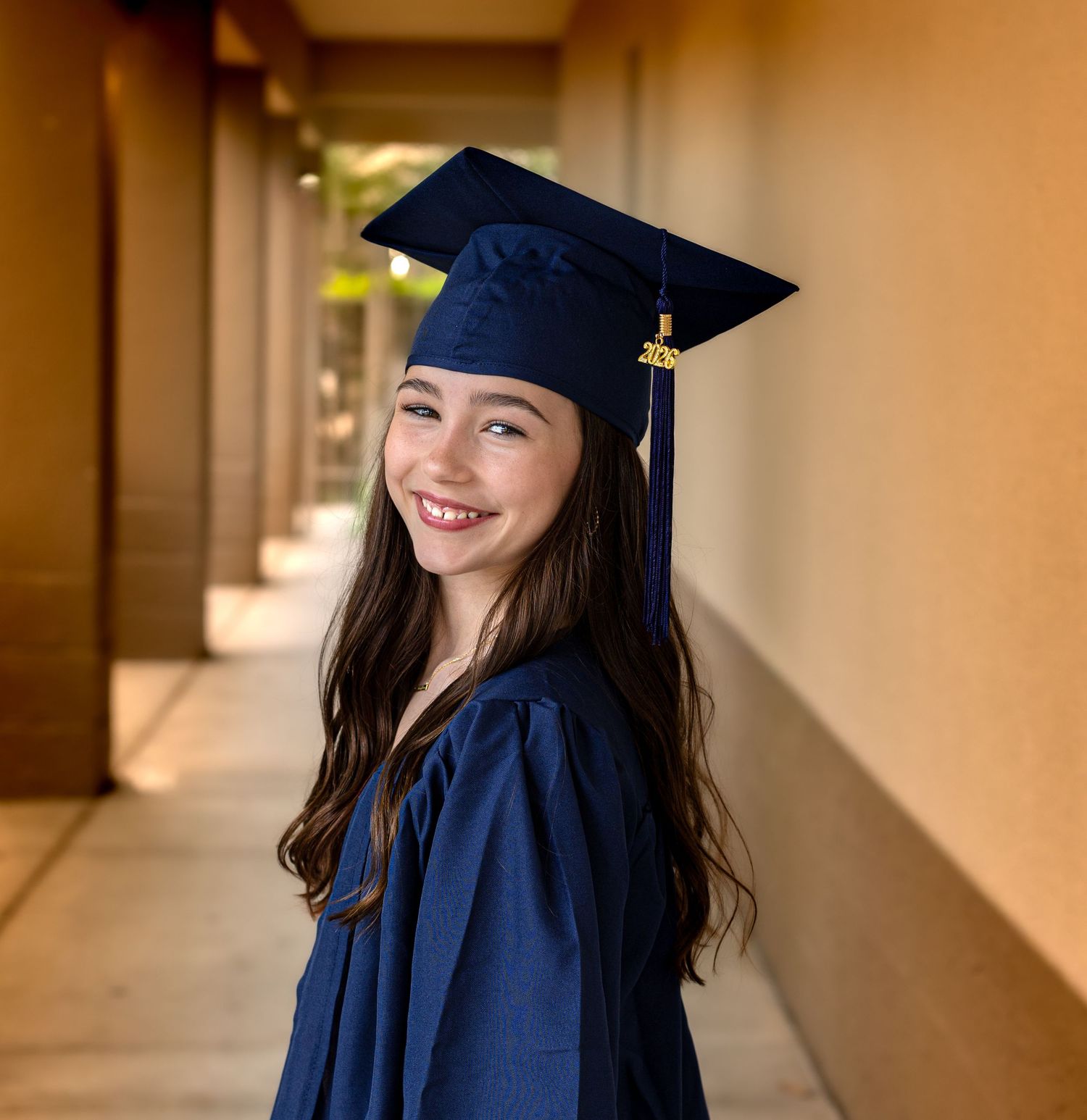 Smiling young woman in navy blue graduation cap and gown posing in a sunlit outdoor corridor.