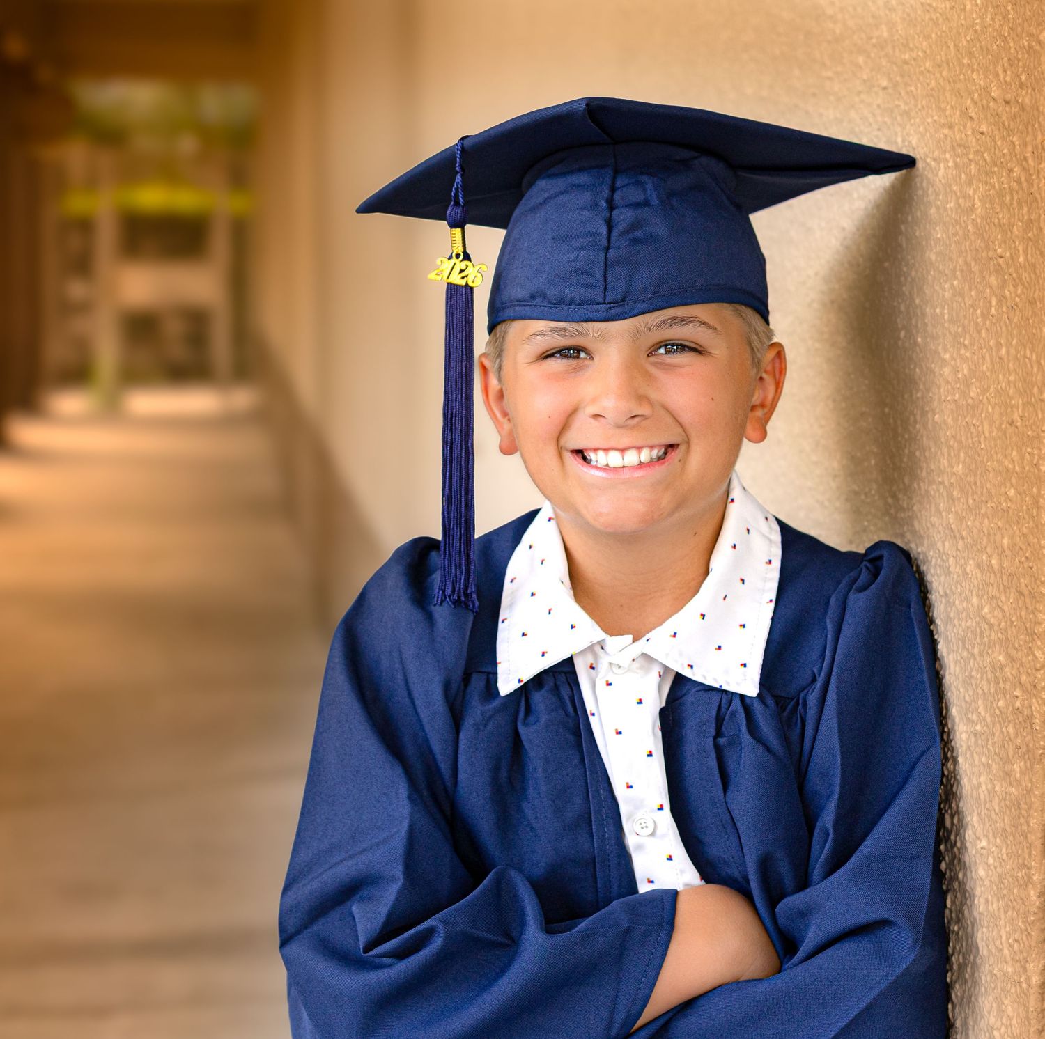 Smiling child in navy blue graduation cap and gown leaning against a wall with arms crossed.