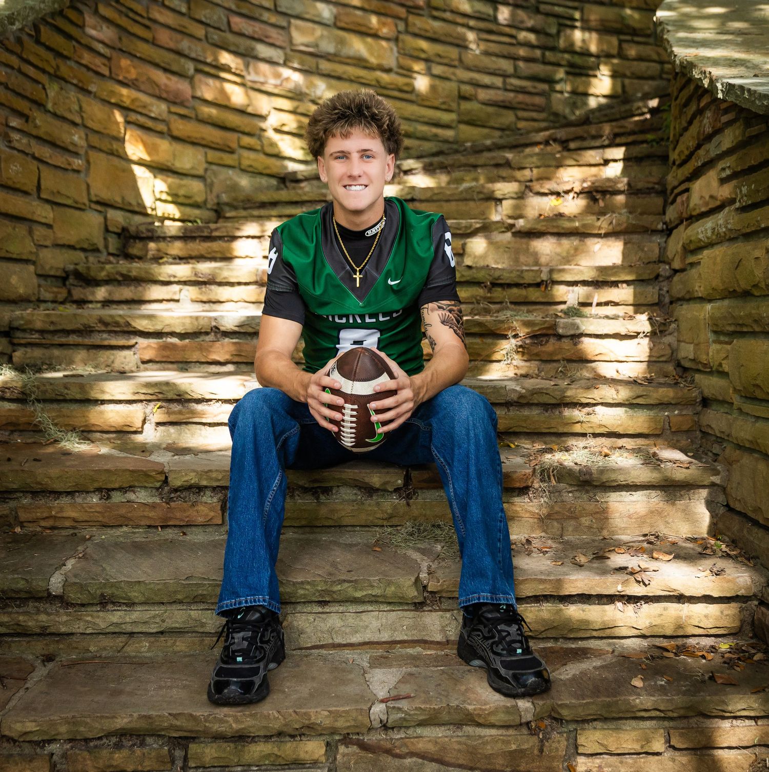Young man in green football jersey sitting on stone steps holding a football, smiling for senior photos.