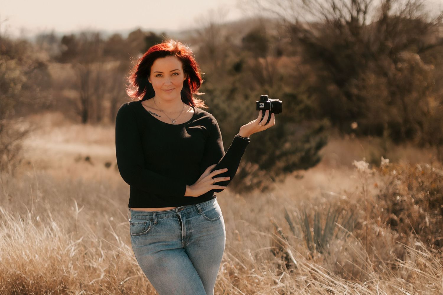 Red-haired woman holding a camera in a golden field, wearing jeans and black top, outdoor photography portrait. Sara Hayes Photography STL