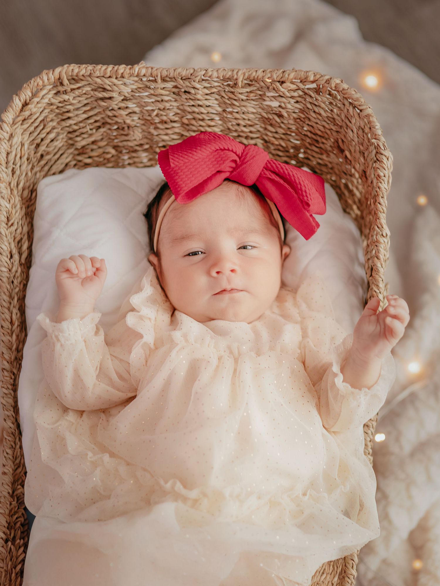 Alert newborn baby girl in lace dress and red bow headband lying in a wicker Moses basket.