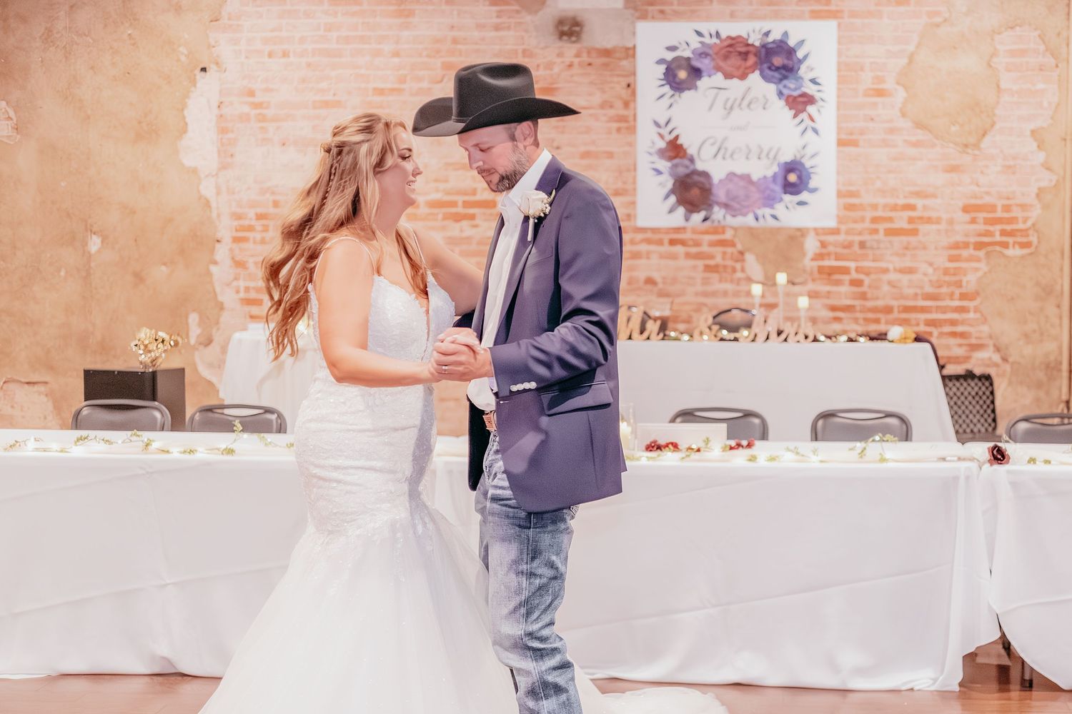 Bride and groom share first dance, groom wearing black cowboy hat and purple blazer in rustic brick reception hall.