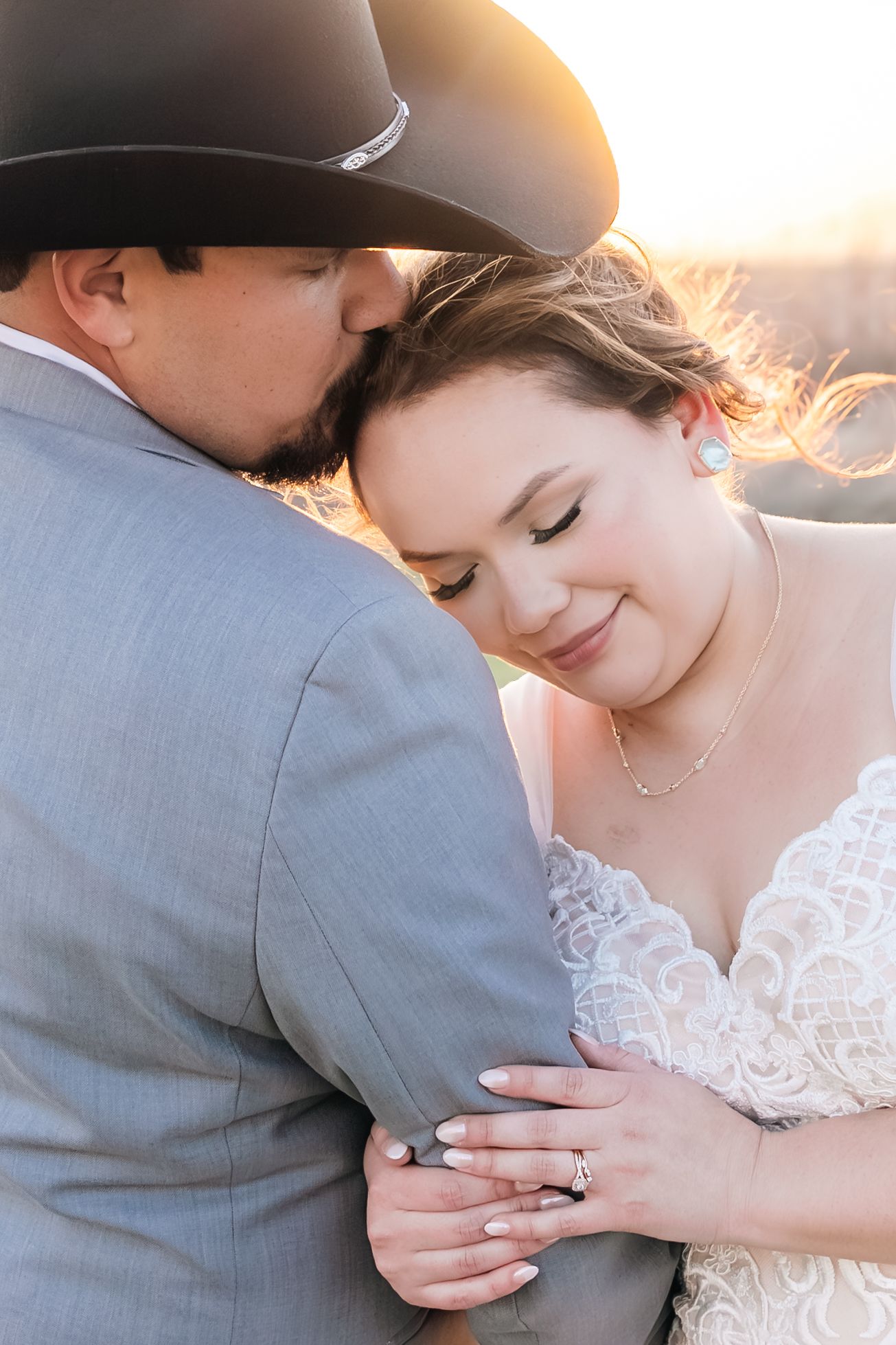 Groom in black cowboy hat kisses bride on the forehead as she smiles warmly during golden hour portraits.