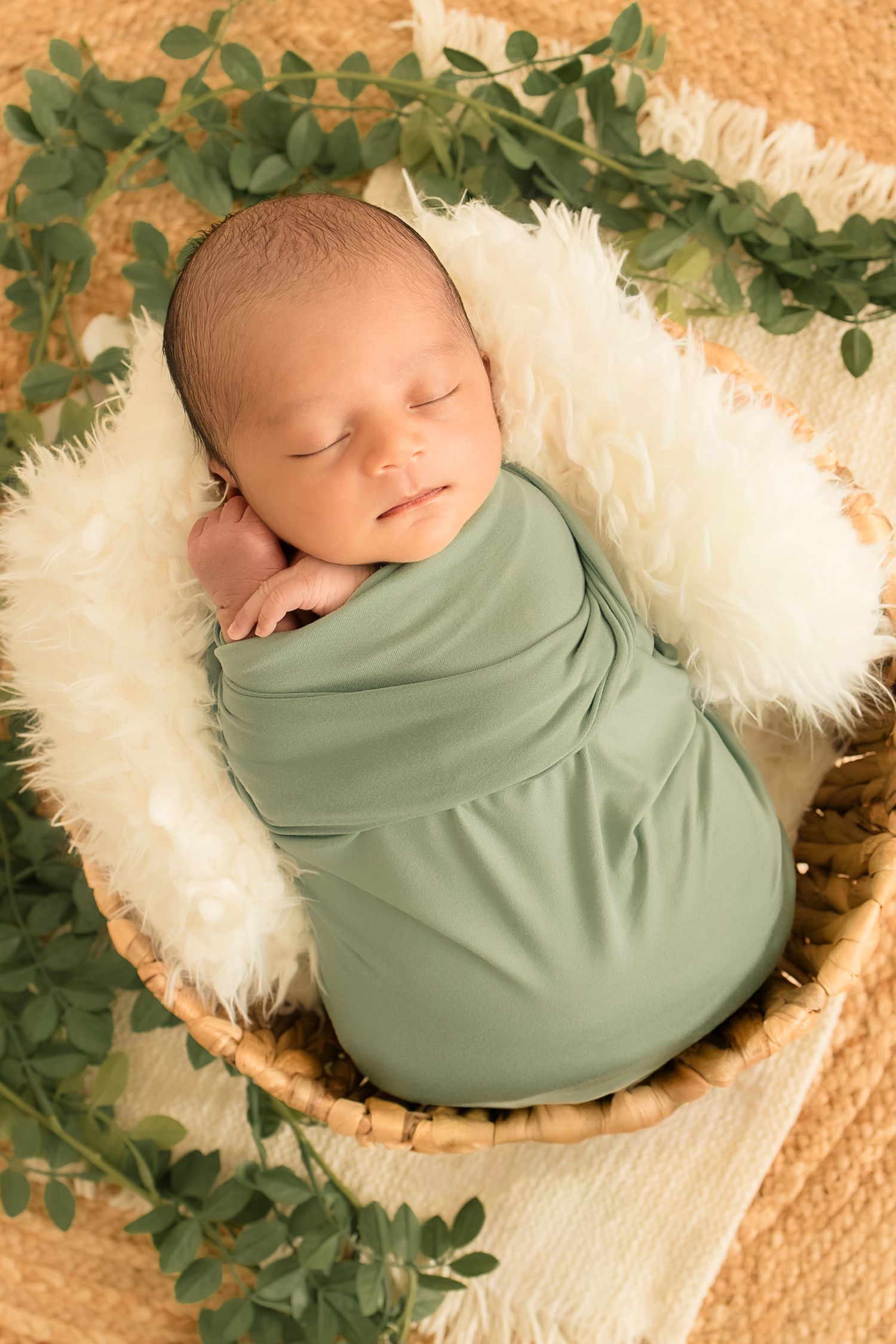 Sleeping newborn baby swaddled in sage green wrap, nestled in a wicker basket with white fur and green eucalyptus leaves.