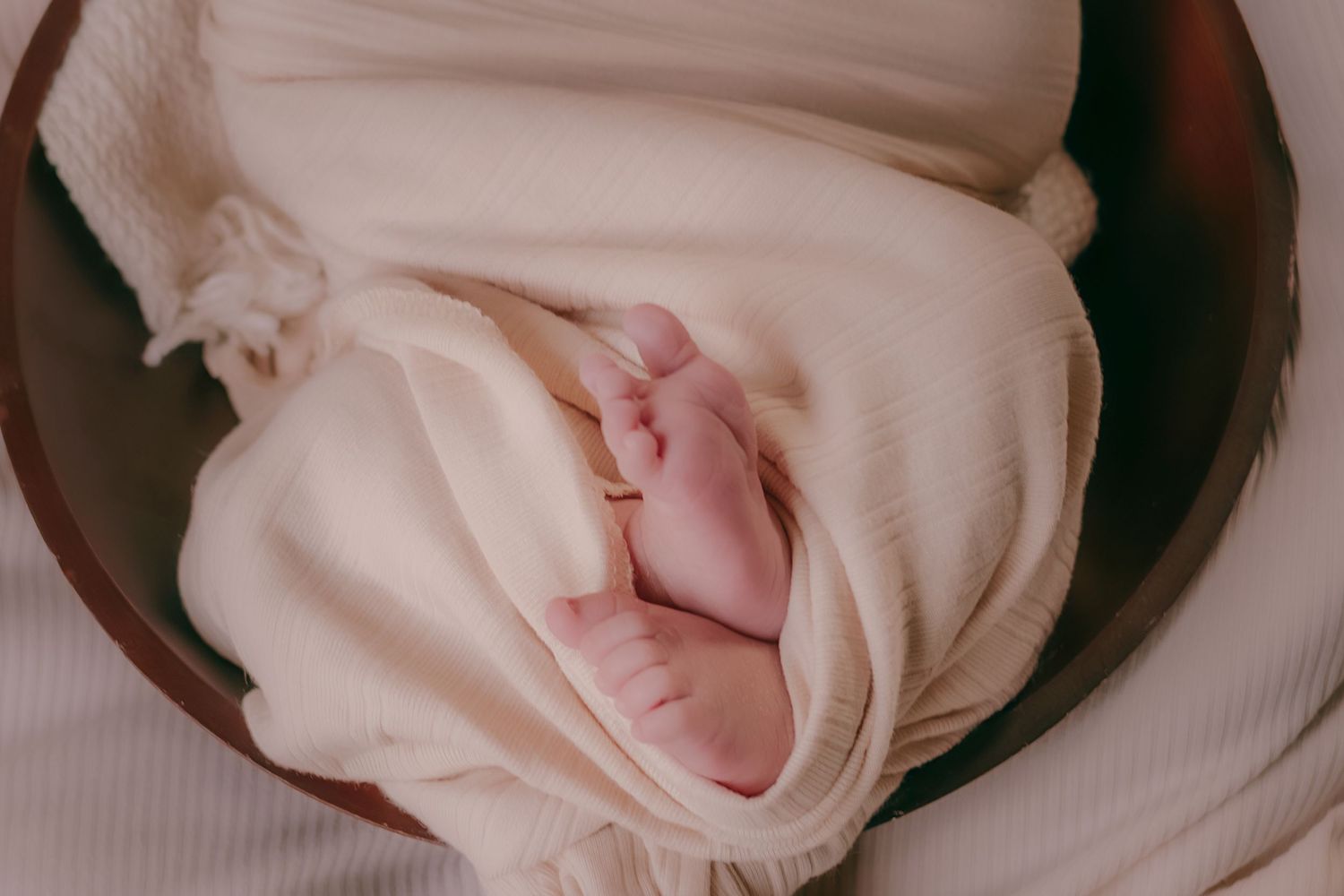 Close-up of newborn baby tiny feet peeking from cream swaddle wrap in wooden bowl.