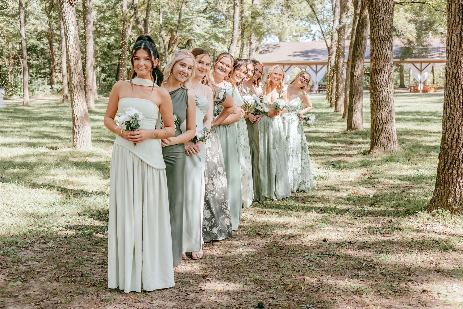 Bridesmaids in sage green dresses hold white bouquets while posing in a line among trees.