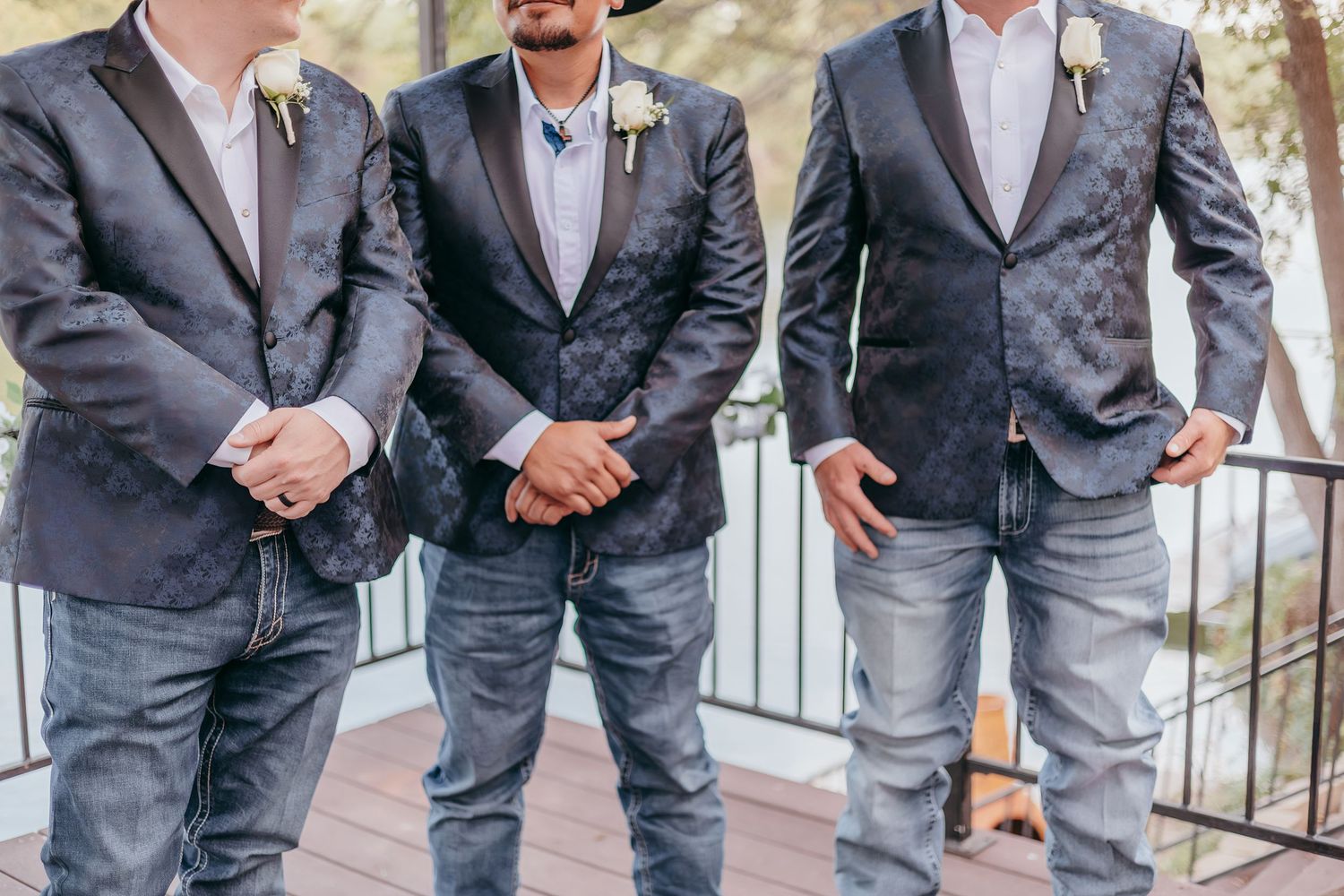 Three groomsmen in matching dark patterned blazers and jeans stand together at outdoor wedding.