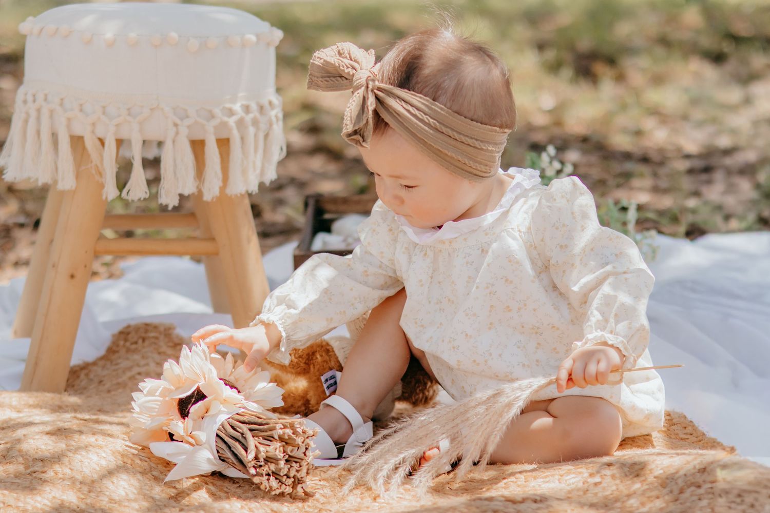 Toddler girl in floral dress and bow headband playing with toys on a blanket outdoors in warm sunlight.