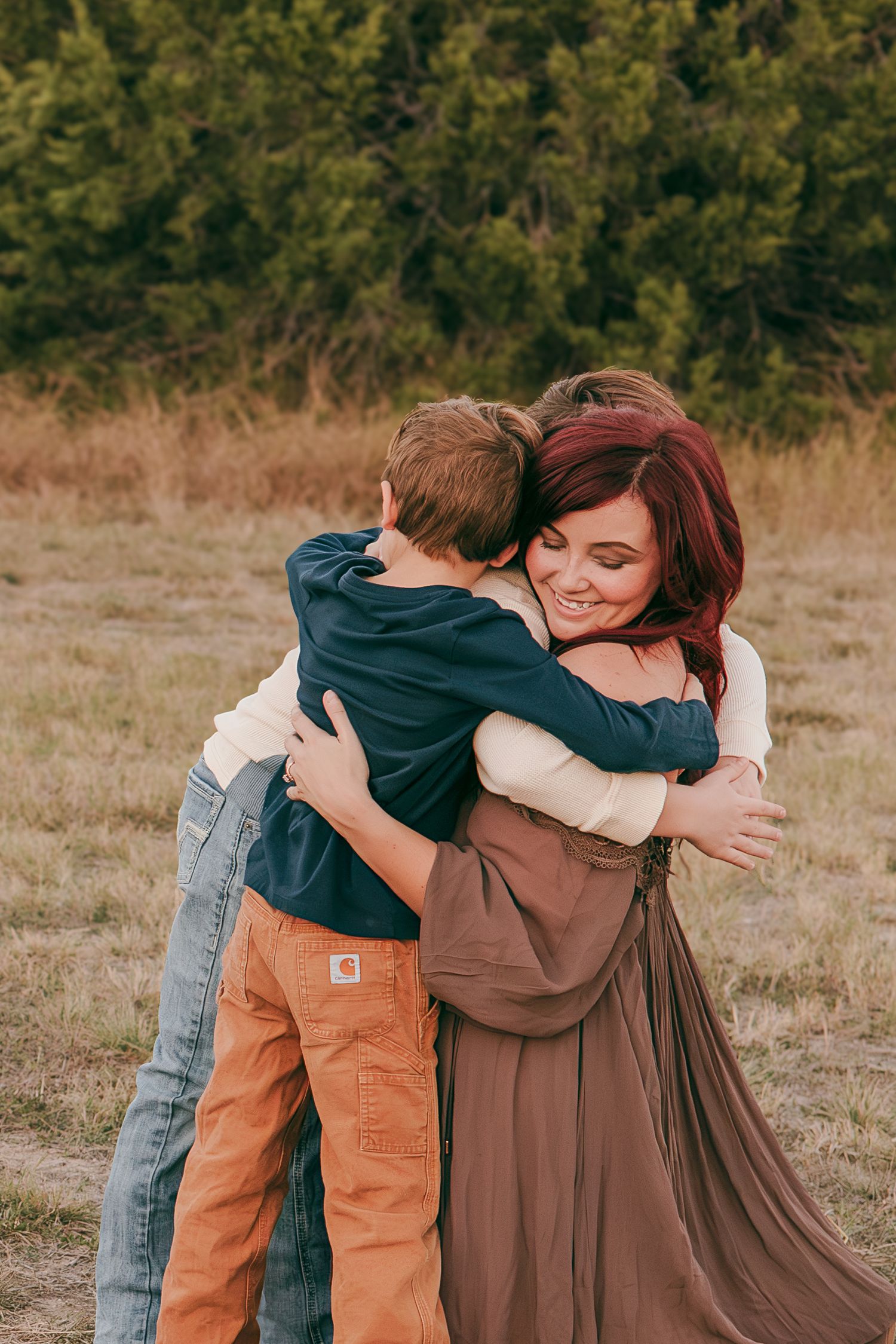 A woman with red hair smiles while hugging two young boys in a field during an outdoor family photo session.