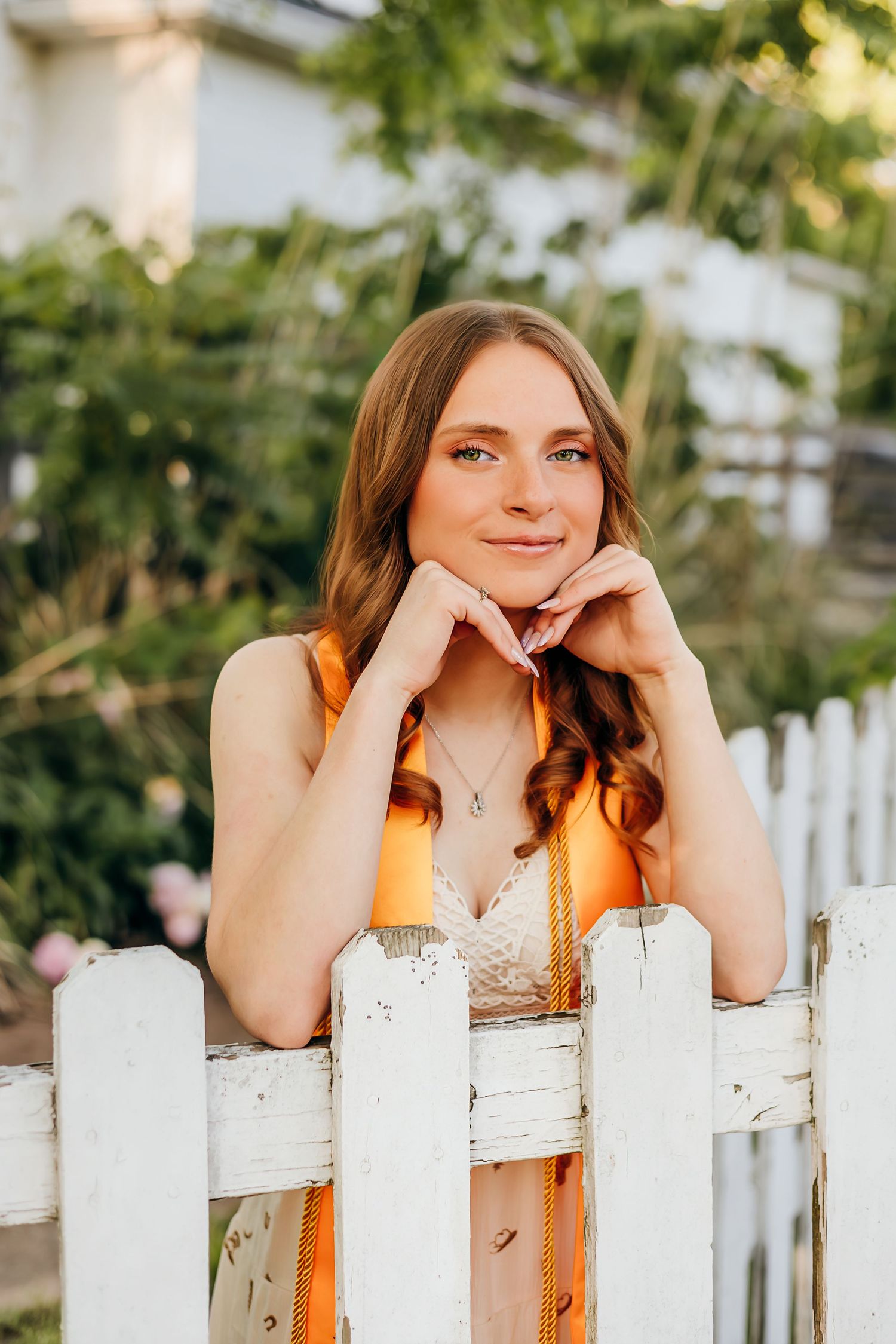 Young woman in orange graduation sash leans on white picket fence, smiling outdoors with greenery behind her.
