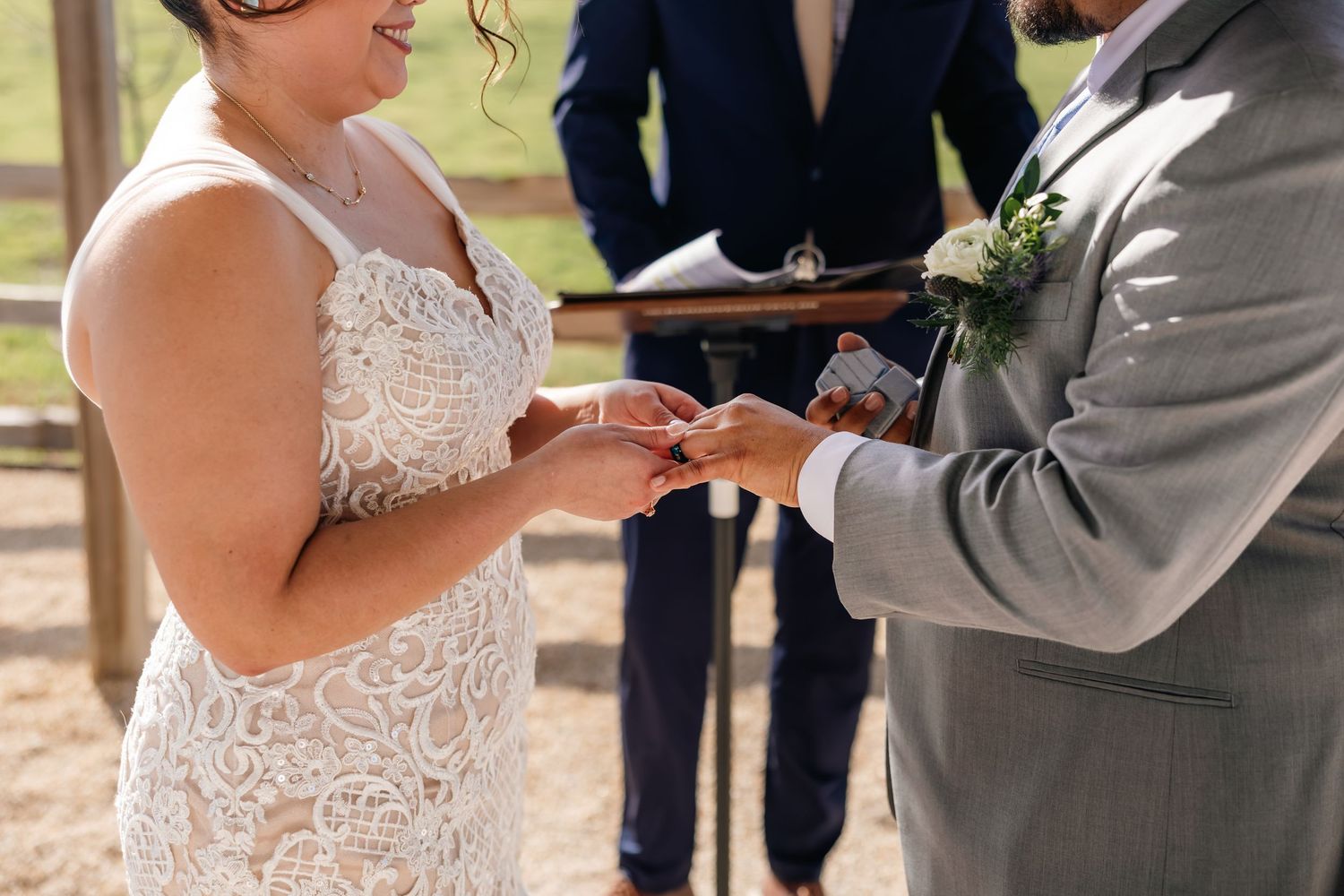 Bride in lace wedding dress placing ring on groom's finger during outdoor wedding ceremony.