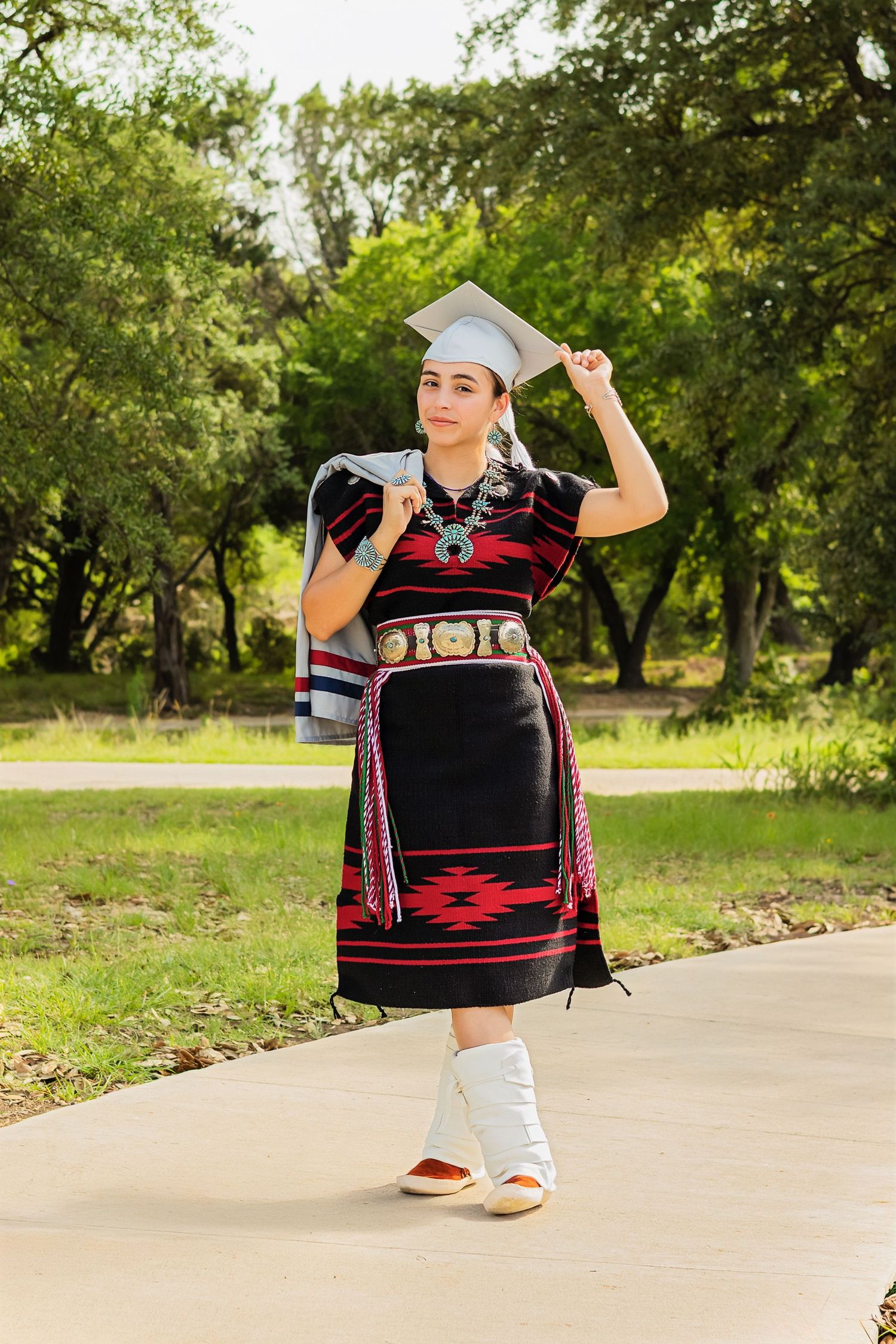 Young woman in traditional Native American dress and graduation cap poses outdoors on a sunny day.