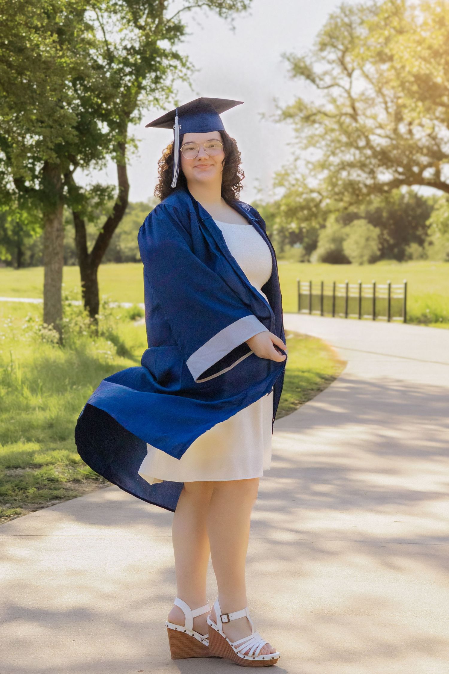 Young woman in blue graduation cap and gown poses outdoors on sunny day, holding diploma in white dress and wedge sandals.
