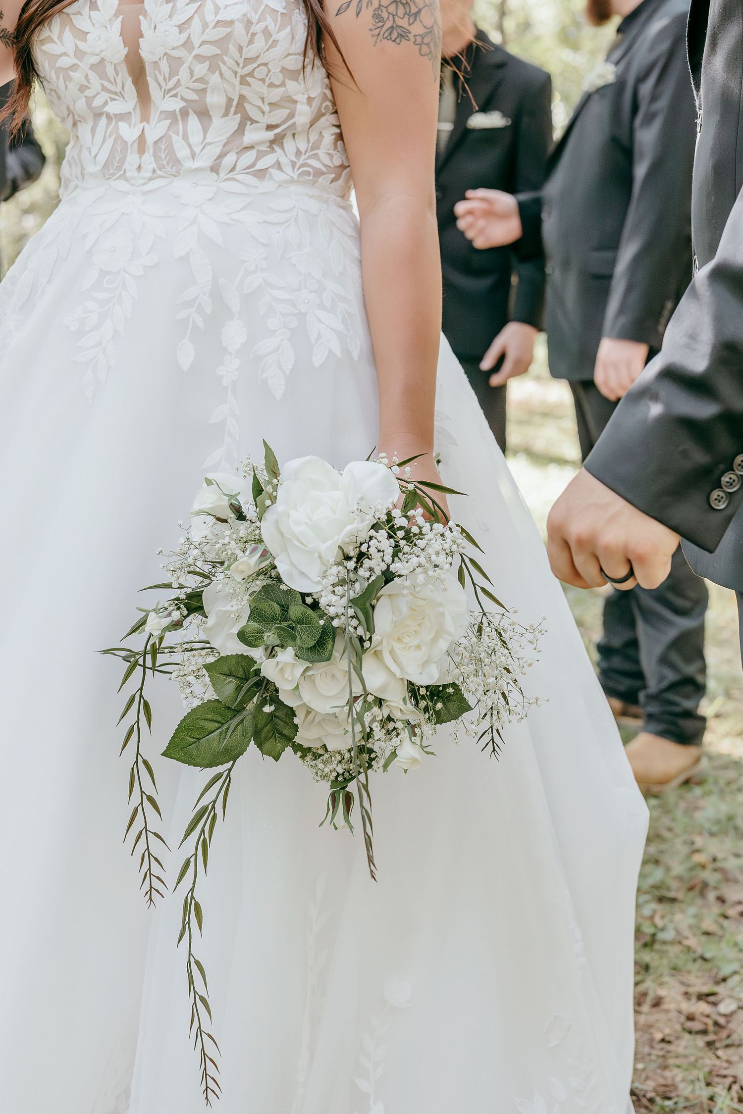 A bride in a lace wedding dress holds a white rose and greenery bouquet while holding hands with the groom.