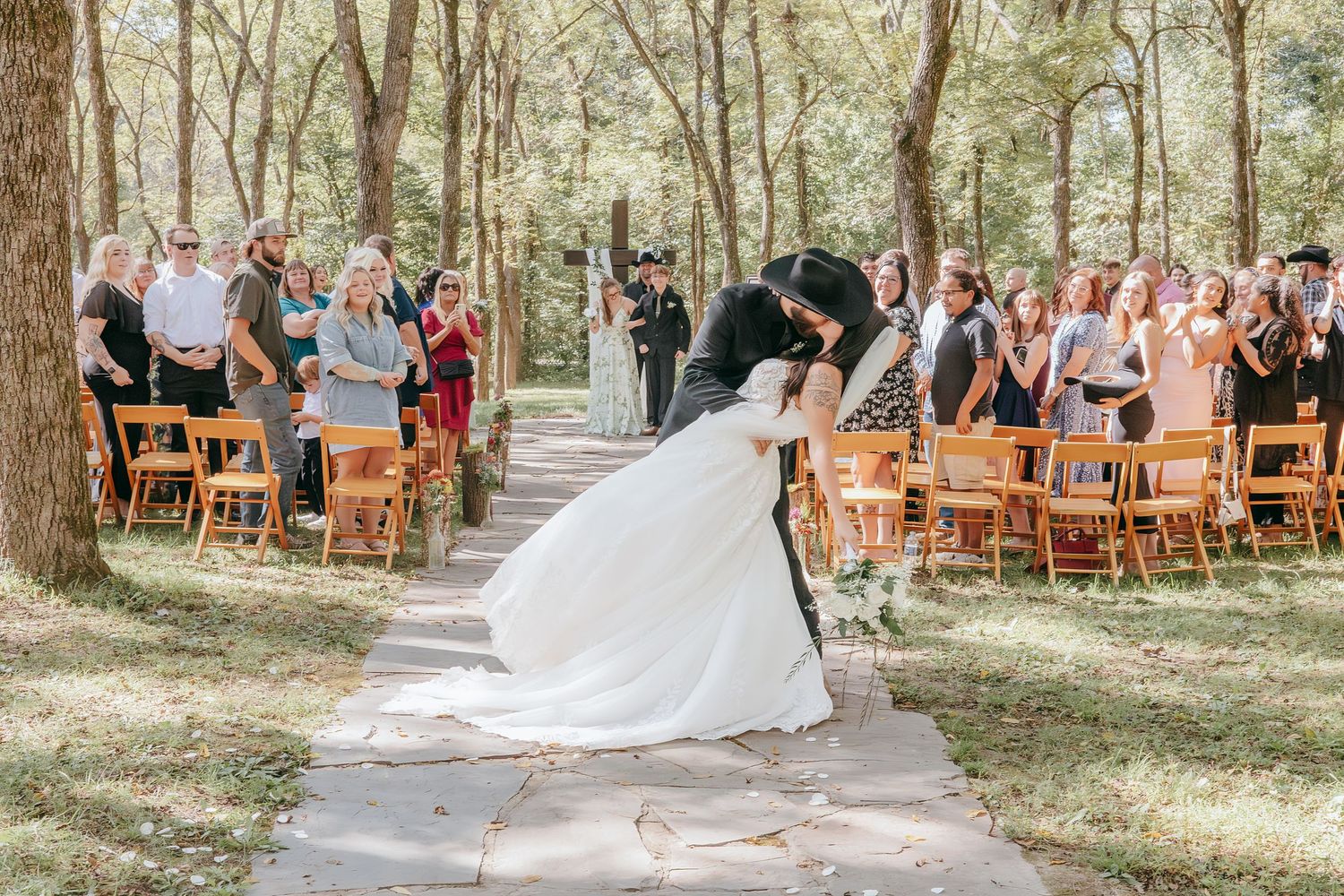 Bride and groom share a first kiss at outdoor woodland wedding ceremony with guests cheering.