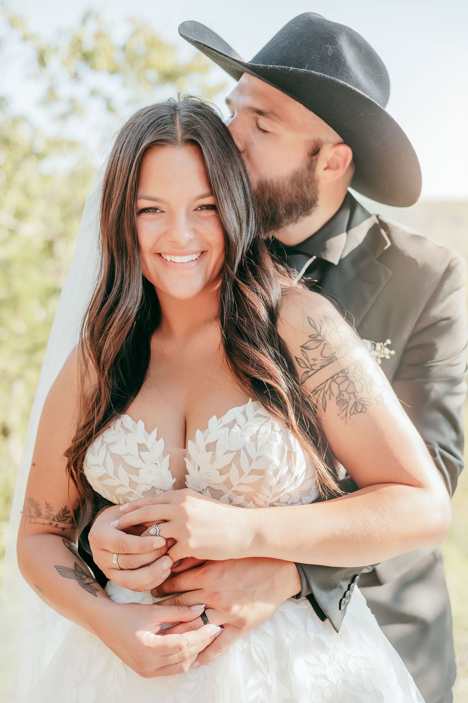 Bearded groom in black cowboy hat kissing bride's cheek as she smiles in floral lace wedding gown.