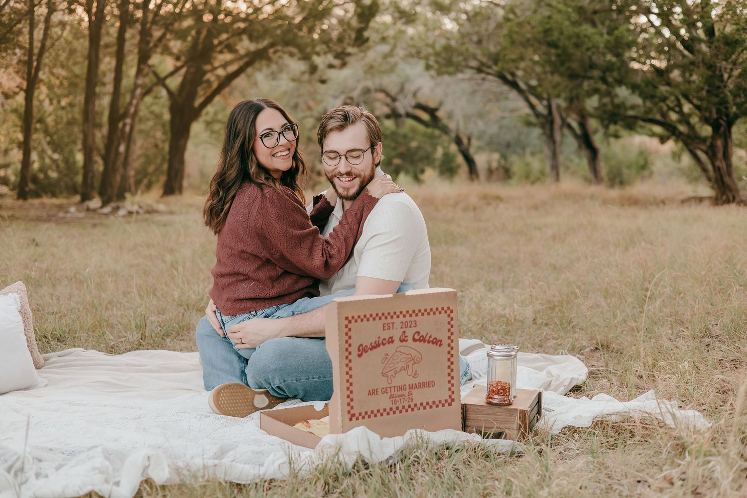 Smiling couple sitting on a white blanket in a field holding a personalized wooden sign.