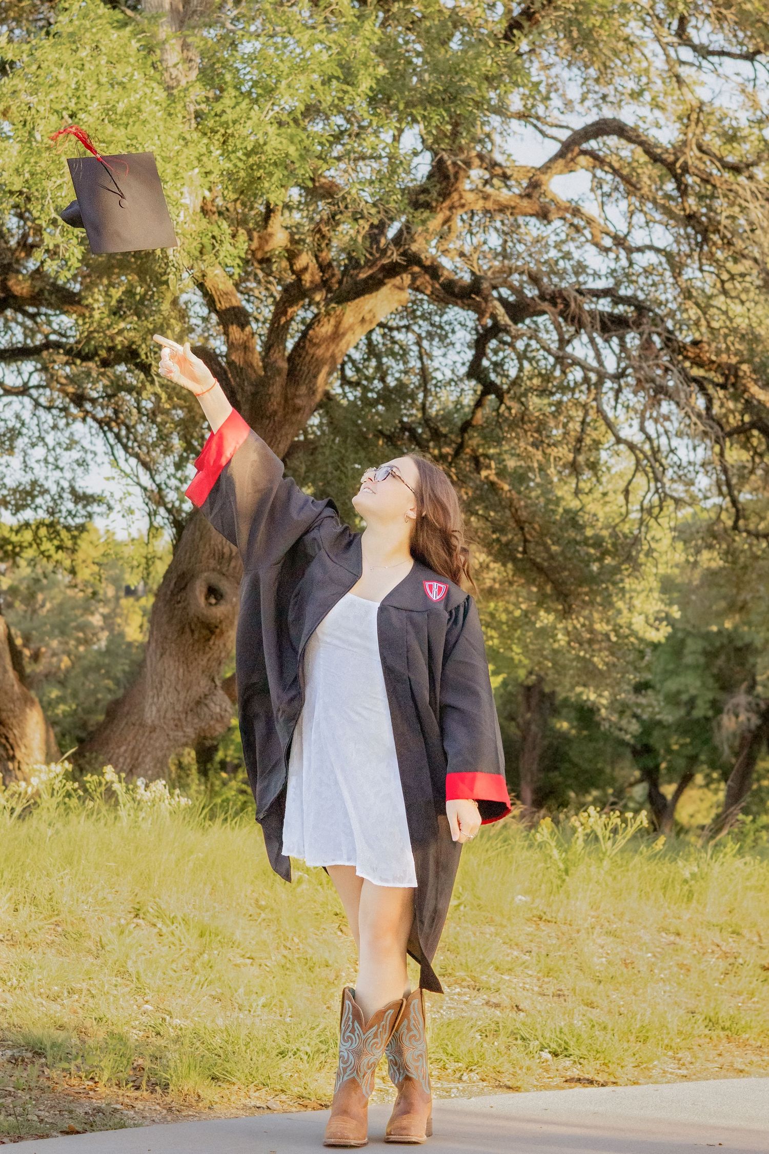 Young woman in graduation gown and cowboy boots tosses cap in air outdoors among trees.