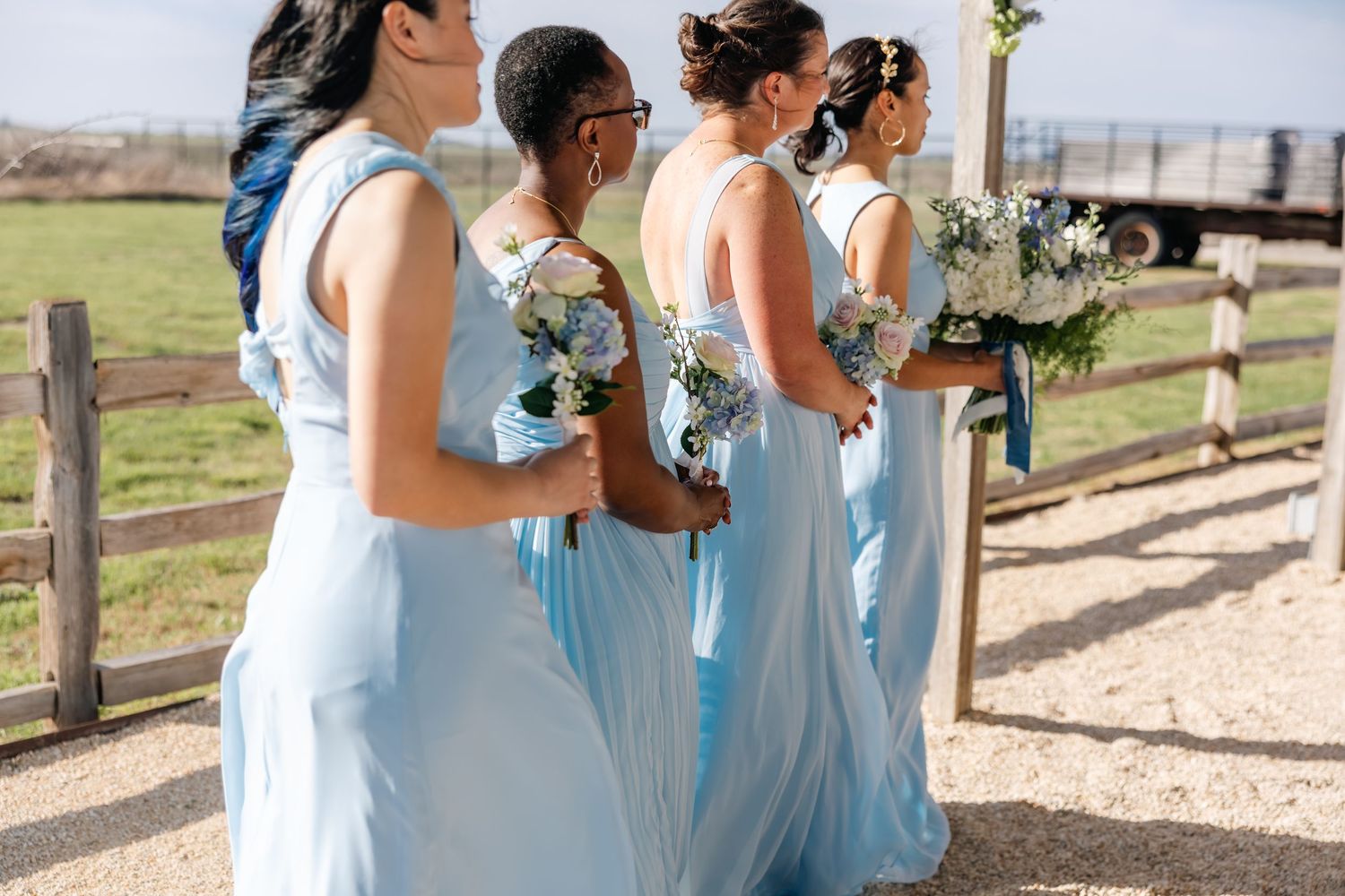 Bridesmaids in light blue gowns holding white floral bouquets stand by a wooden fence at an outdoor wedding.