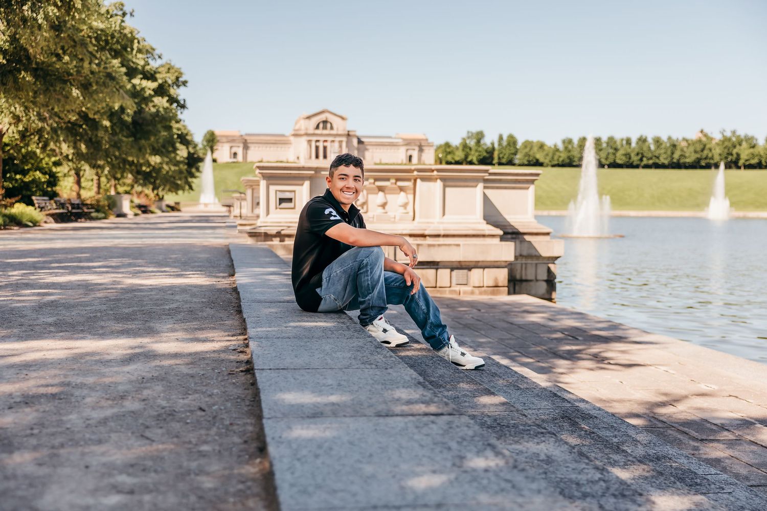 Young man sitting on stone steps by a fountain and lake near a classical building on a sunny day.