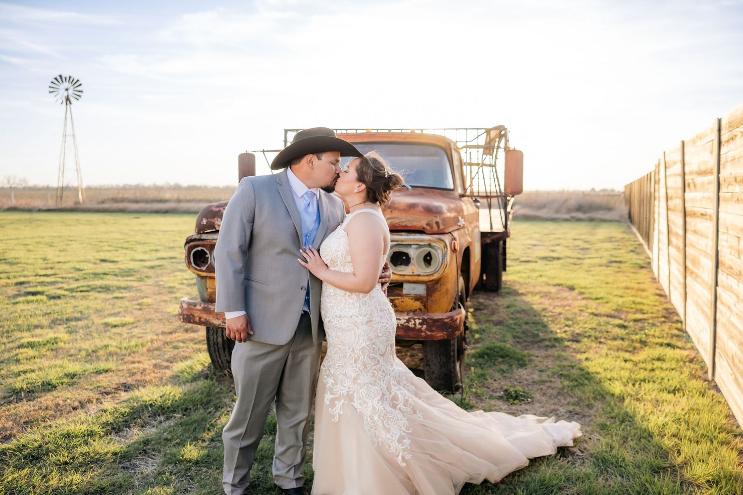 Bride and groom share a kiss in front of a vintage rusty truck at a rustic farm wedding at golden hour.