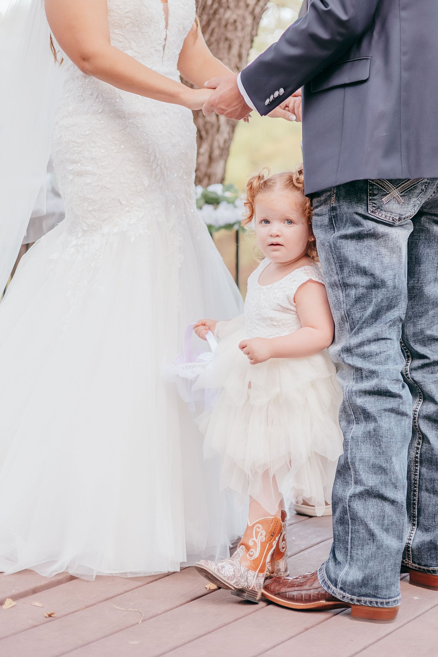 A flower girl in a white tutu dress and cowboy boots stands between a bride and groom holding hands at a wedding.