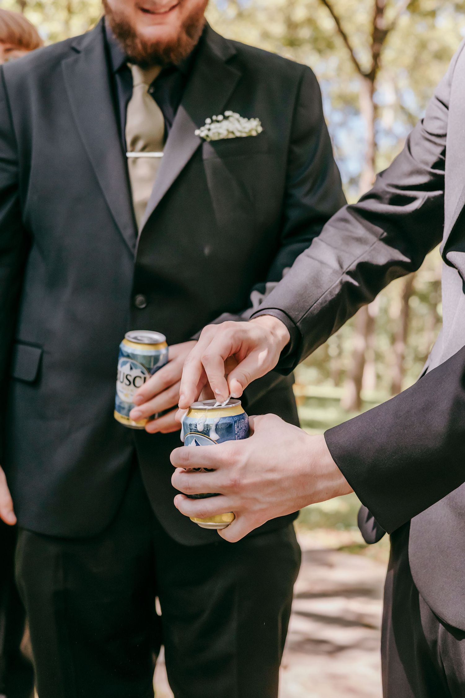 Groomsmen in dark suits opening Busch beer cans outdoors during wedding celebration in wooded setting.