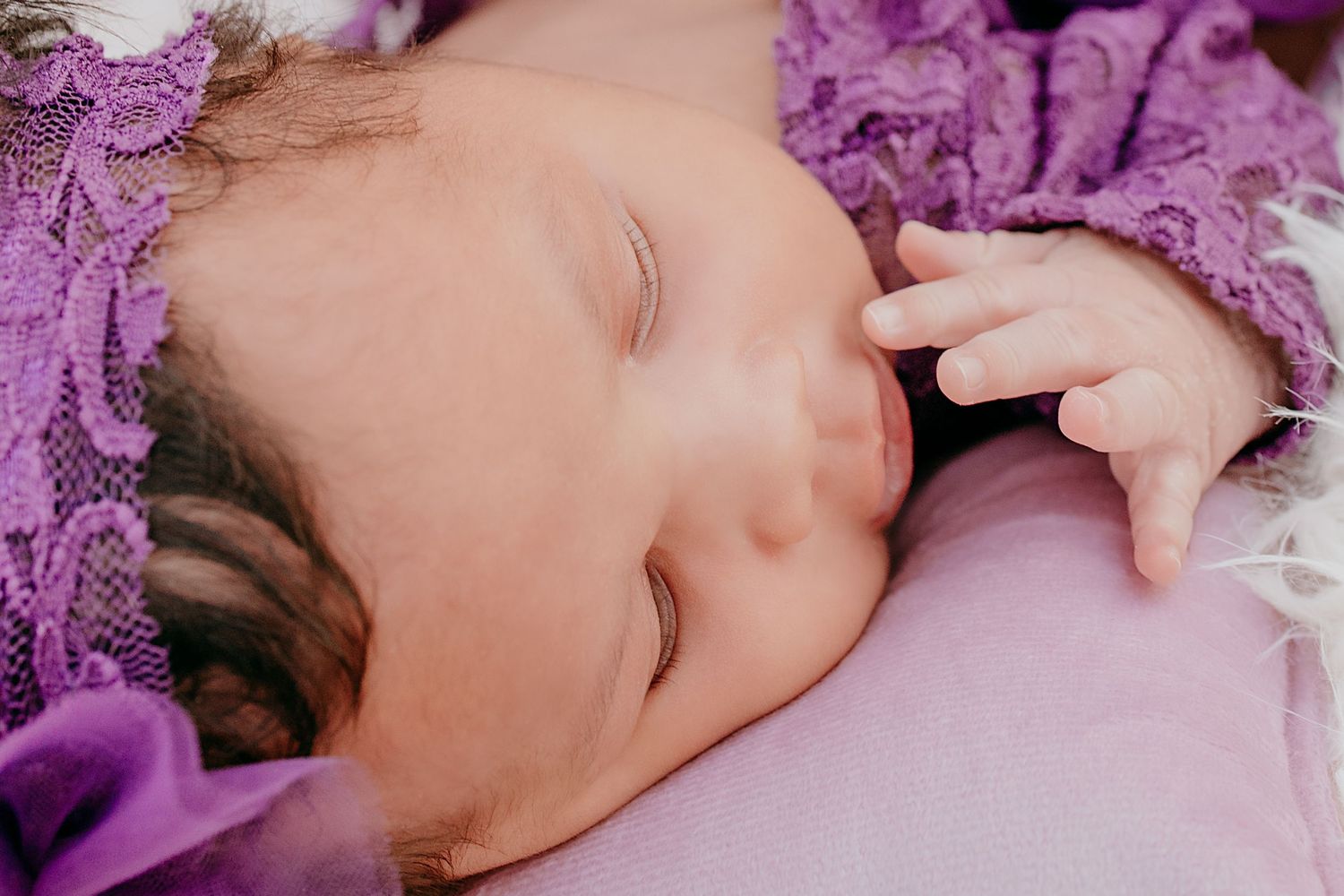 Close-up of peaceful sleeping newborn baby wearing purple lace outfit and floral headband.