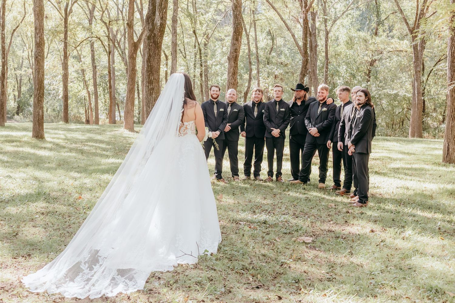 Bride in white lace gown and veil facing groomsmen lined up in dark suits in sunlit forest clearing.
