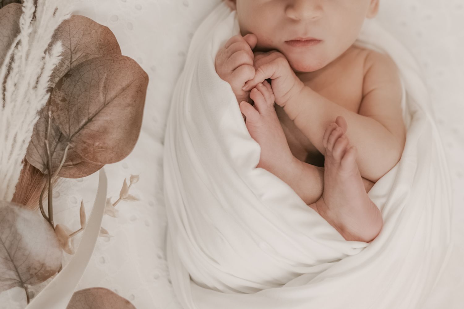 A newborn baby rests in a white swaddle surrounded by dried botanicals in a soft, neutral setting.