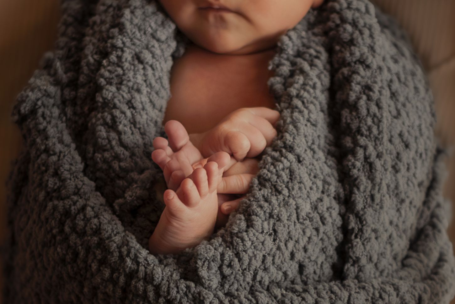 Newborn baby wrapped snugly in a soft gray knit blanket, with tiny hands visible in a peaceful pose.