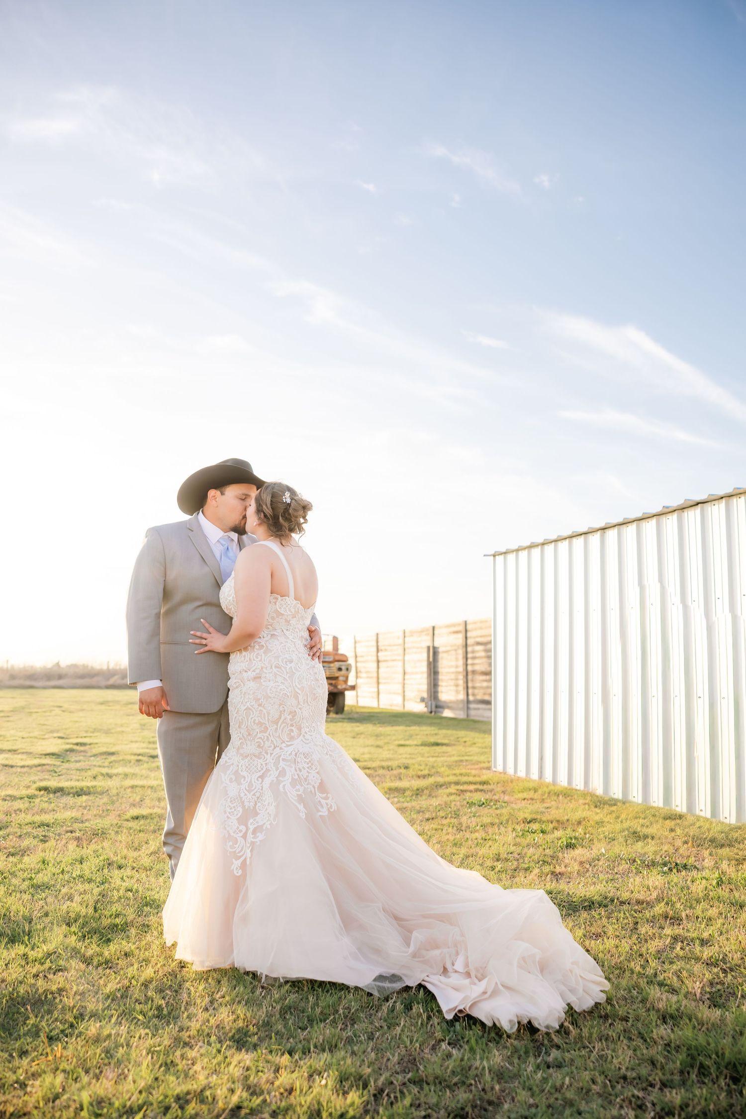 Bride and groom share a romantic kiss at golden hour on a farm, bride wearing a lace mermaid gown with a train.