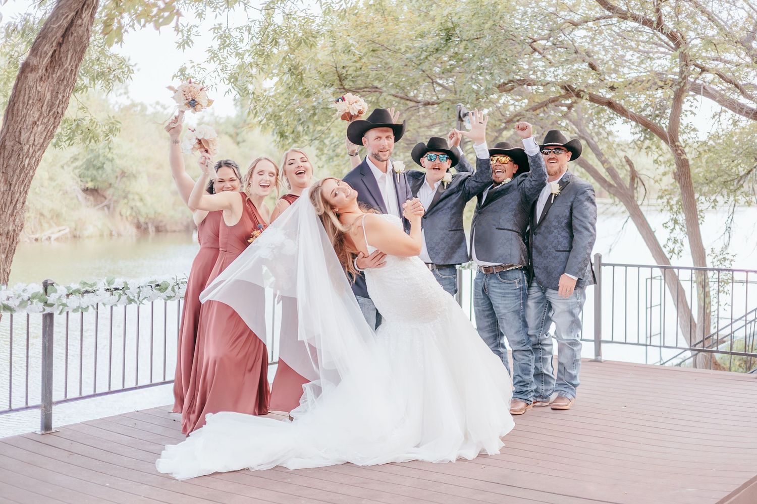 Wedding party celebrating on a deck, bridesmaids in pink dresses and groomsmen in cowboy hats near a river.