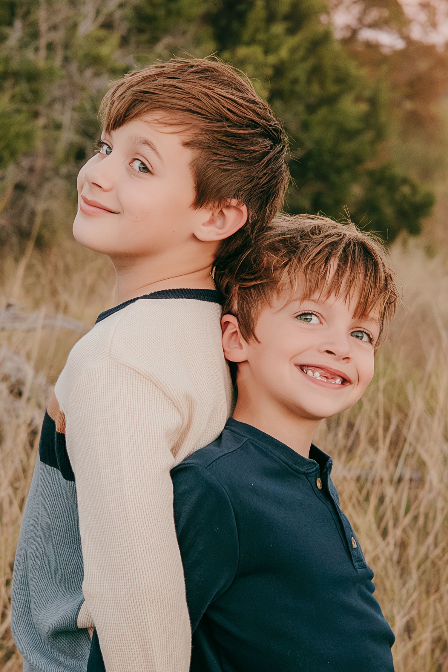 Two young brothers smile back-to-back in a field of tall golden grass during an outdoor portrait session.
