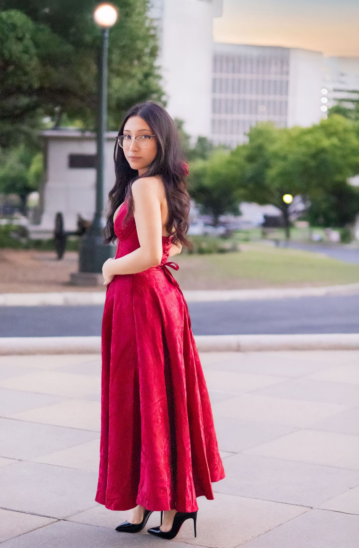Woman in elegant red gown and heels posing outdoors in urban park setting at dusk.