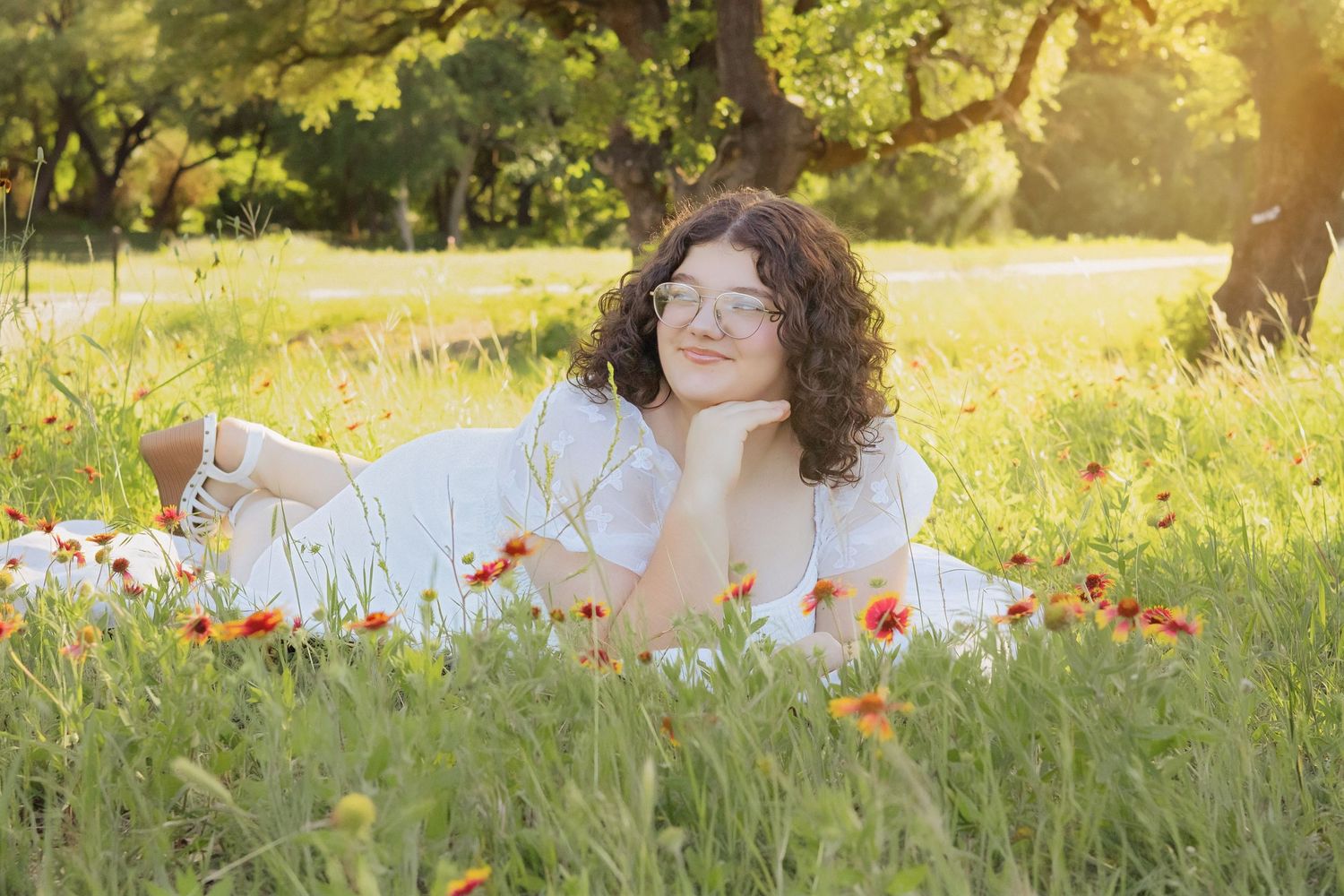 Young woman with curly hair and glasses in white dress lying in sunlit wildflower meadow with red blooms.