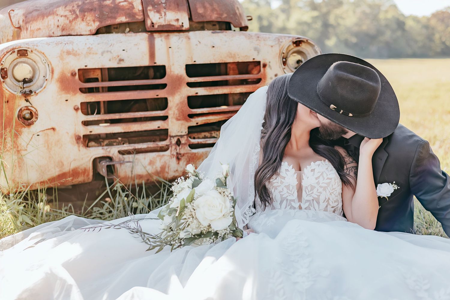 Bride and groom kissing beside a rustic vintage tractor in a field, groom wearing a black cowboy hat. St Clair Missouri