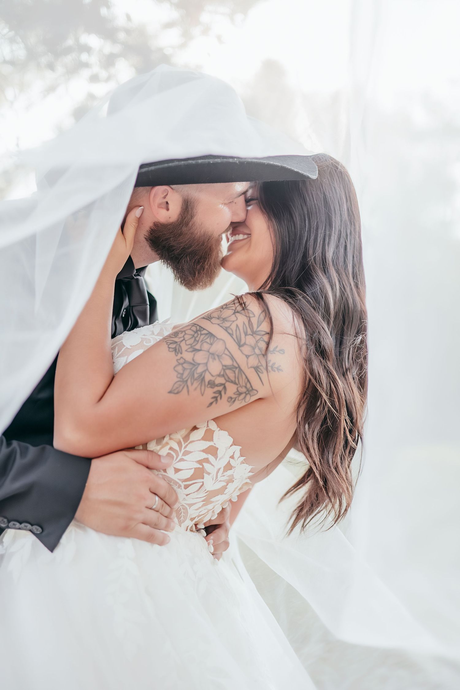 Bride and groom sharing romantic kiss under flowing white veil, bride with floral arm tattoo visible.