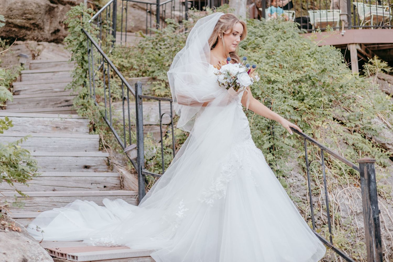Elegant bride in a flowing white ball gown and veil descending rustic wooden stairs outdoors.