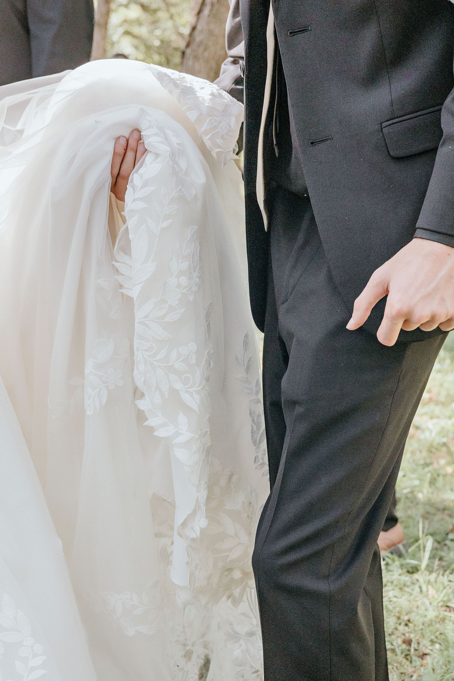 A bride in a lace wedding gown stands beside a groom in a black suit at an outdoor wedding.