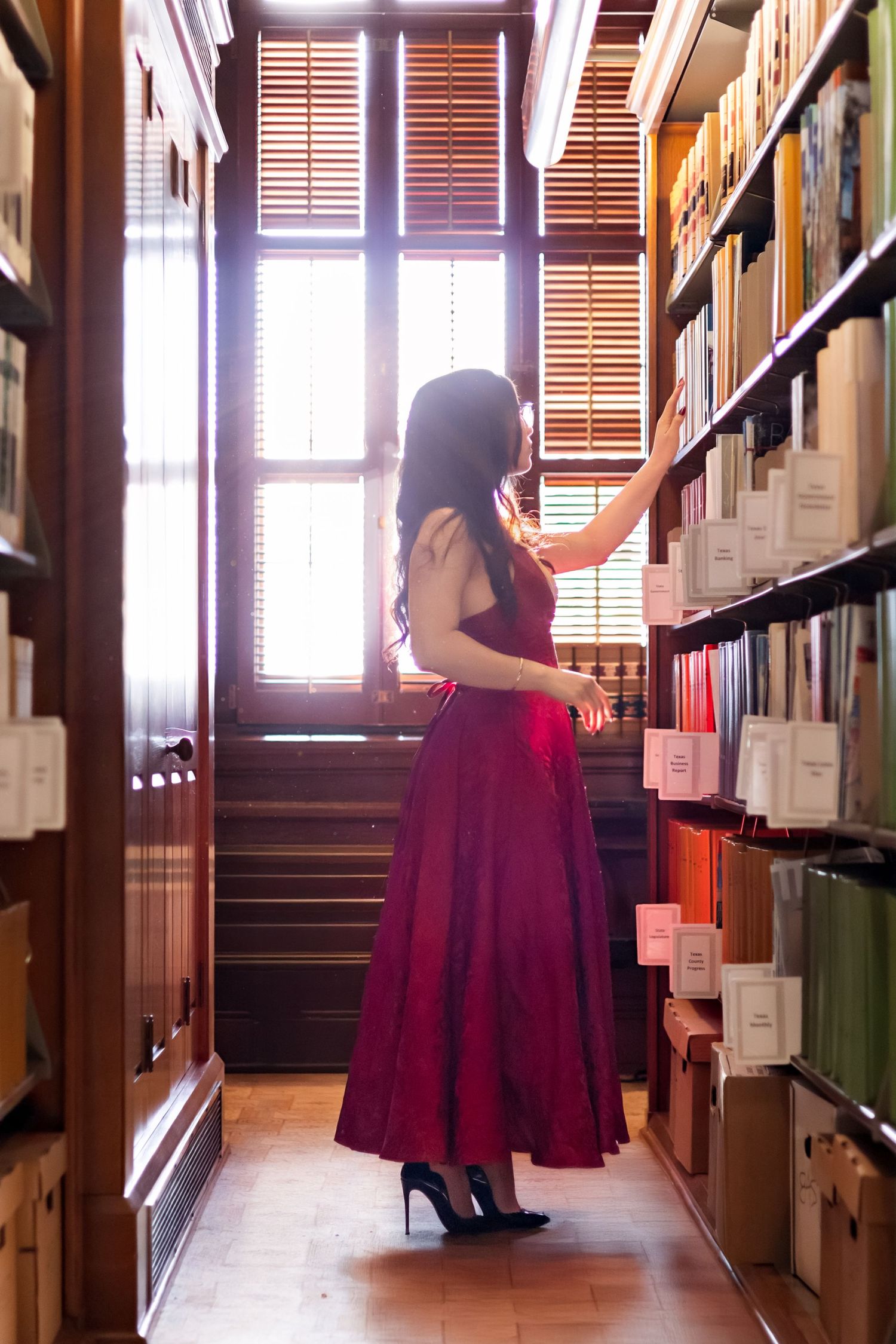 Young woman in red dress browses books in sunlit library aisle between tall shelves.