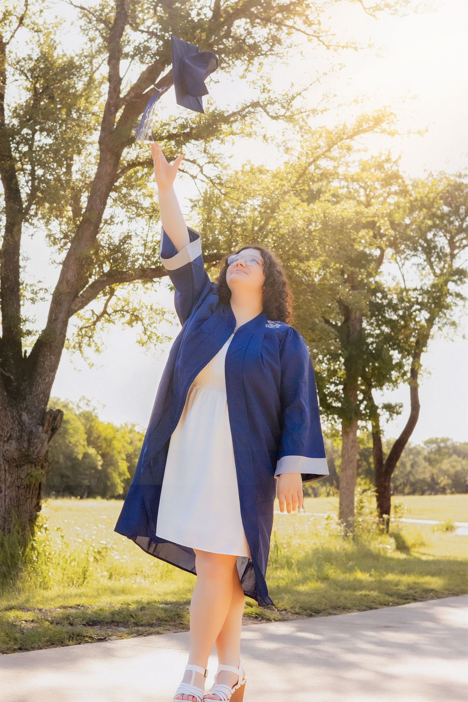 Young woman in blue graduation gown tossing cap in the air outdoors, celebrating academic achievement among green trees.
