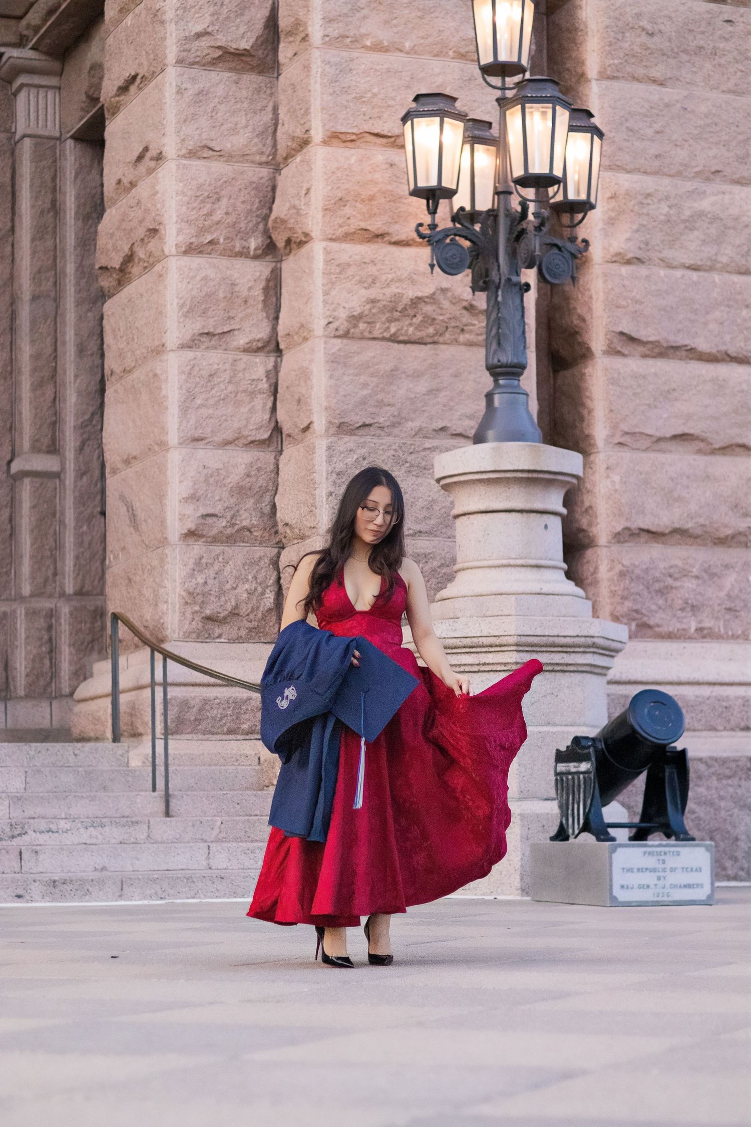Young woman in red gown holding graduation cap outside stone building with ornate lamp post.
