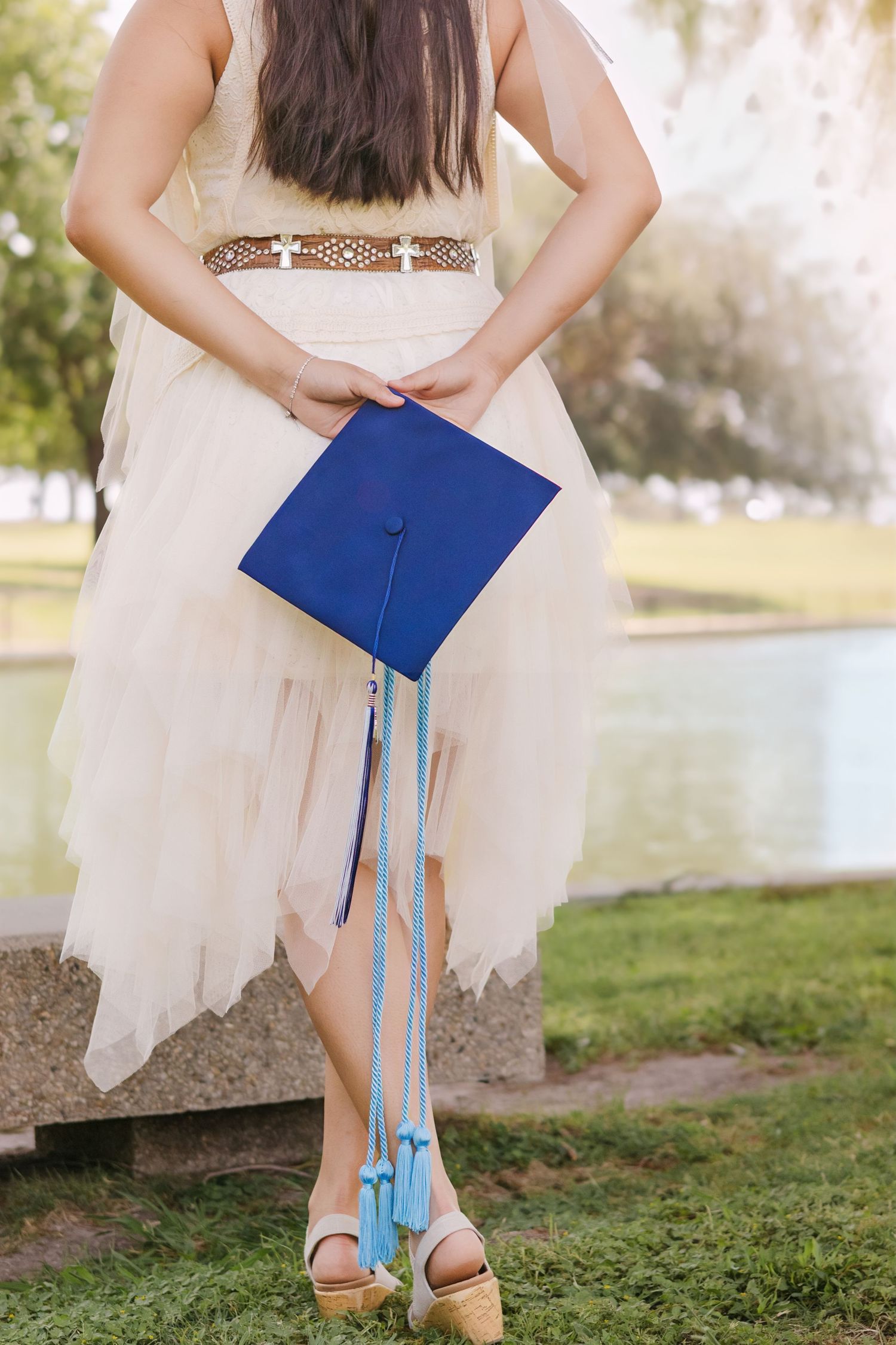 Senior Girl in white dress holding blue graduation cap behind her back outdoors near a pond. St Louis Senior photography