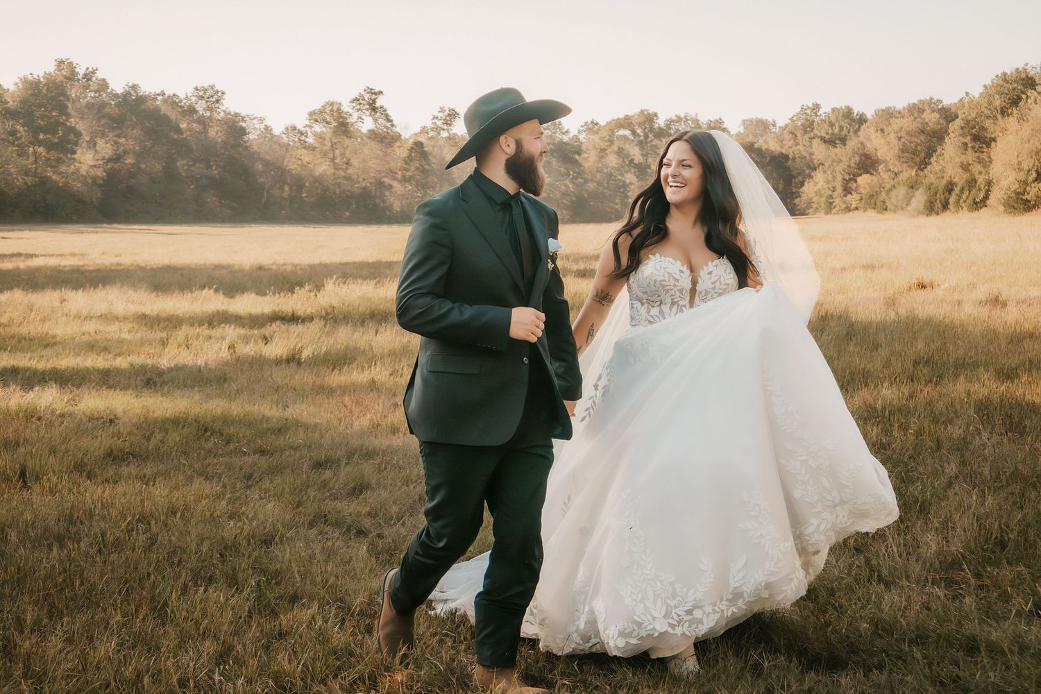 A bride in a lace wedding gown and groom in a green suit walk joyfully through a golden field.
