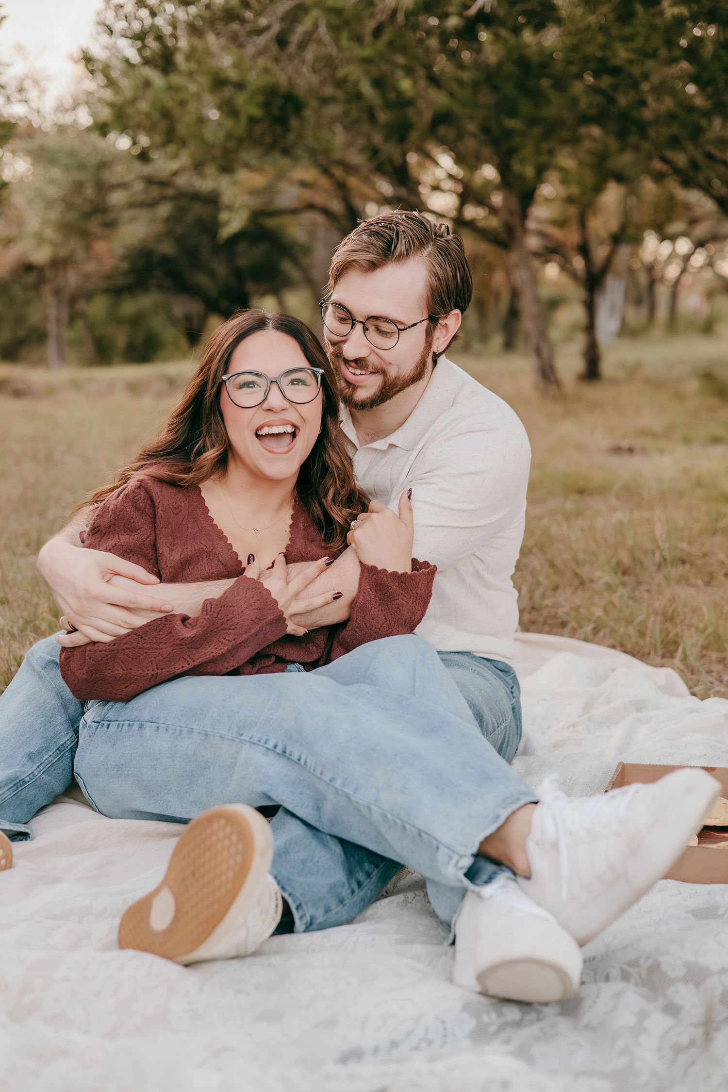 Happy couple sitting on a blanket outdoors laughing and embracing in a green park setting.