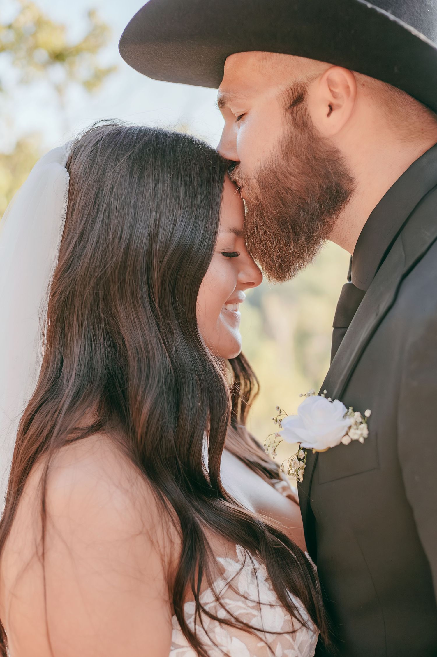 Bride and bearded groom in black cowboy hat sharing a tender forehead-to-forehead moment outdoors.