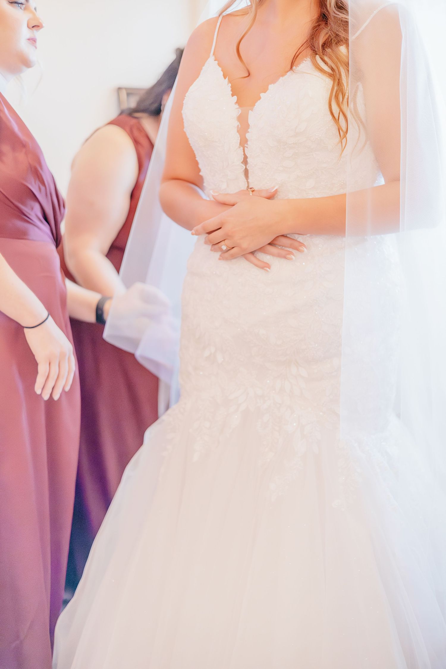 Close-up of a bride in a lace mermaid wedding gown with veil assisted by bridesmaids.