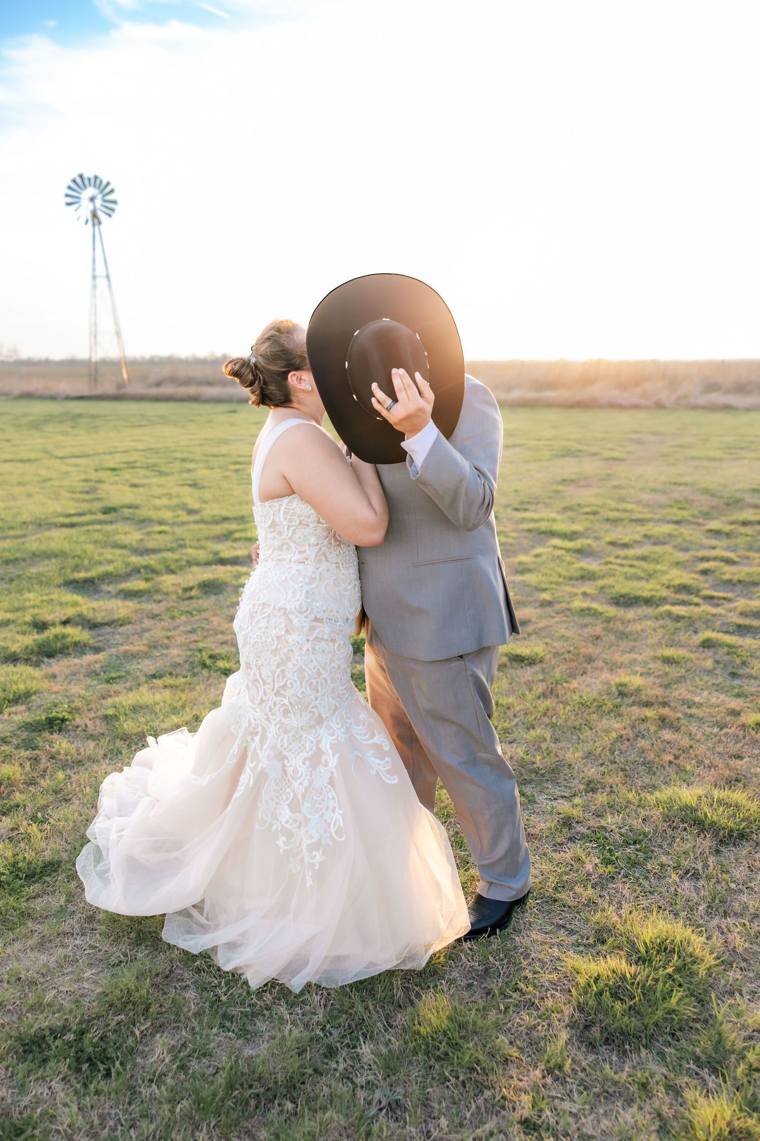 Wedding couple kissing behind a black cowboy hat in a golden sunset field with a windmill in the background.